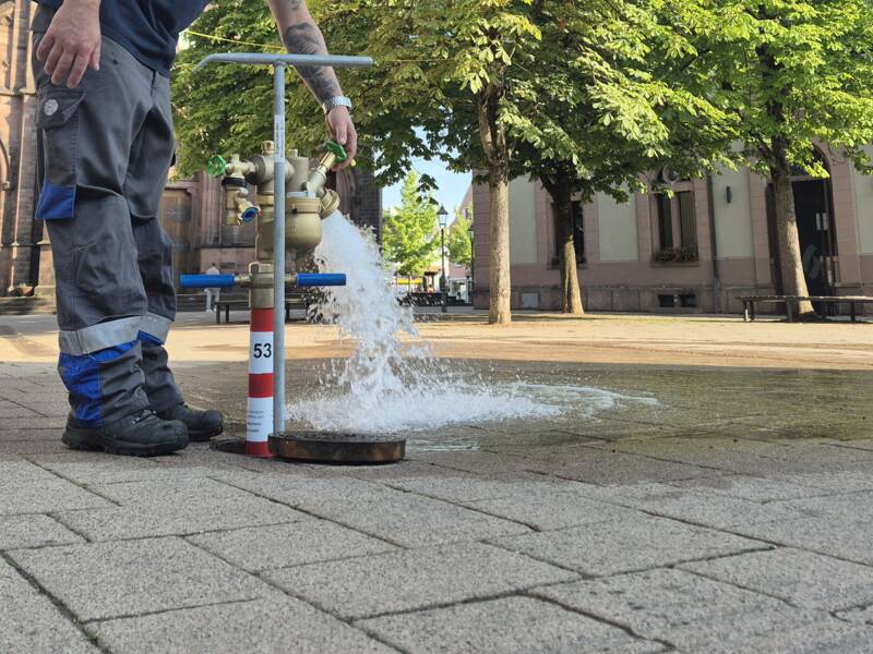 Eine Person dreht einen Hydranten auf einem gepflasterten Platz mit einer Kirche und Bäumen im Hintergrund auf.