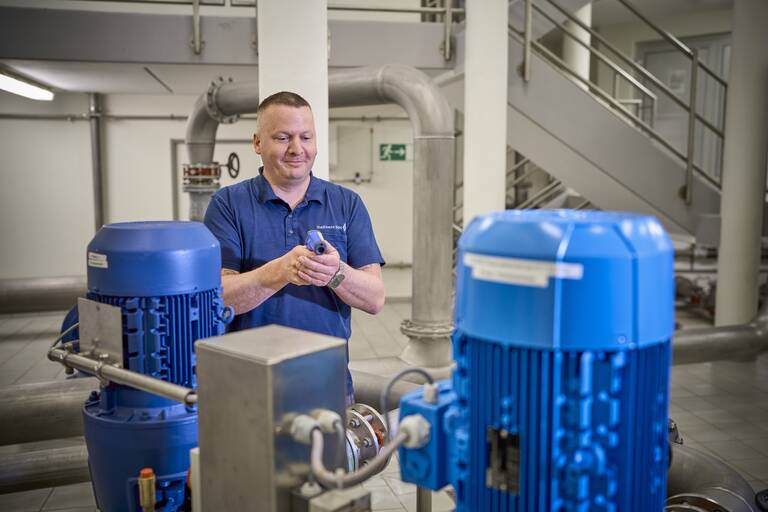 Ein Mann in Arbeitskleidung steht lächelnd in einer industriellen Umgebung zwischen blauen Maschinen und hält ein kleines Gerät in den Händen.