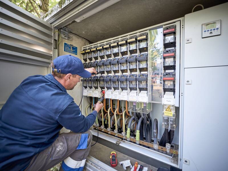 Ein Techniker in blauer Arbeitskleidung arbeitet mit Werkzeugen an einem geöffneten Verteilerkasten voller Schalter und Kabel.