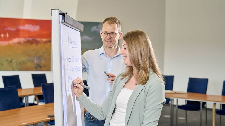 Eine Frau schreibt lächelnd auf ein Flipchart, während ein Mann daneben steht und mit einem Stift auf das Chart zeigt.