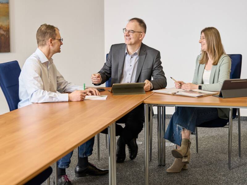 Drei Personen sitzen an einem Tisch in einem Besprechungsraum und unterhalten sich freundlich miteinander.