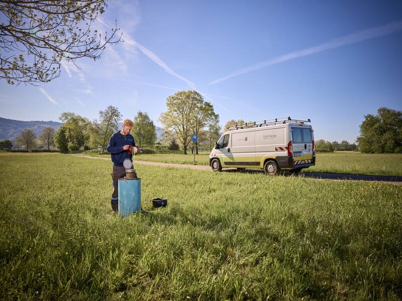 Eine Person arbeitet in einer grünen Wiese an einem Gerät, während ein weißer Transporter mit der Aufschrift "Gute Energie" auf einem Weg in der Nähe geparkt ist.