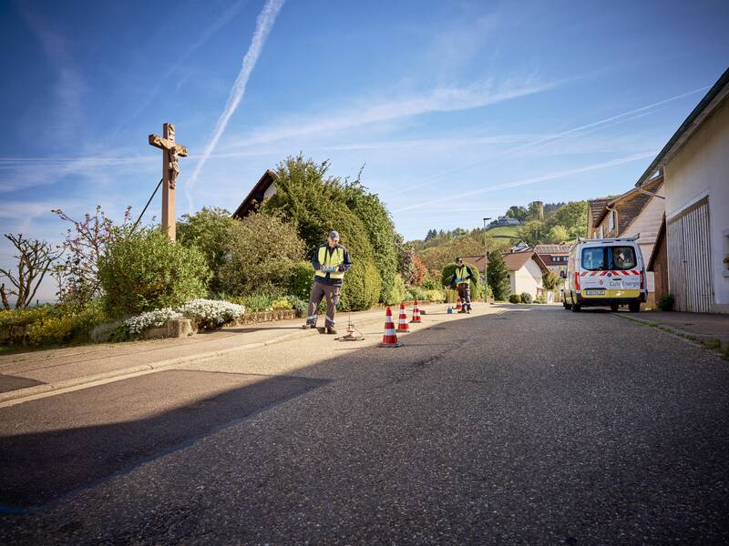 Zwei Wassermonteure in Warnwesten führen Messungen auf einer Straße durch, auf der Verkehrsleitkegel aufgestellt sind, während im Hintergrund eine Landschaft mit Häusern und im die Burg Windeck zu sehen ist.