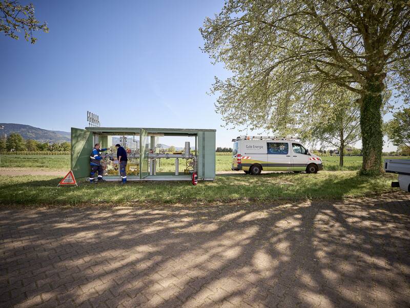 Zwei Personen in Arbeitskleidung arbeiten an einer technischen Anlage auf einer Wiese neben einem weißen Lieferwagen unter einem großen Baum.