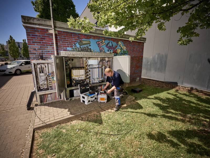 Ein Techniker arbeitet im Freien vor einem geöffneten Verteilerkasten an der Seite eines Backsteingebäudes mit bemalter Wand.