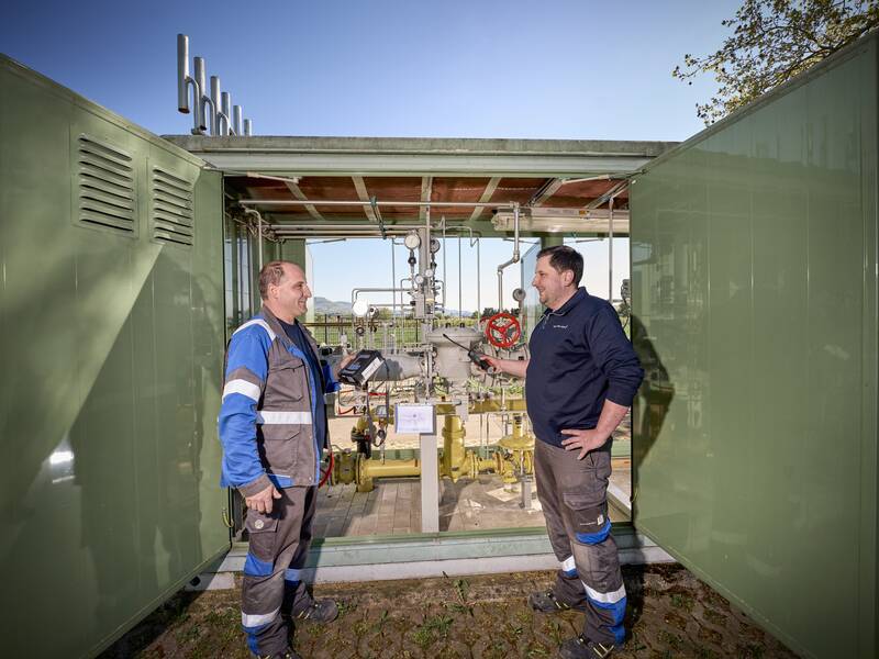 Zwei Männer stehen lächelnd vor einer technischen Anlage mit verschiedenen Rohren und Anzeigeinstrumenten, umgeben von einem grünen Außengelände und einem blauen Himmel im Hintergrund.