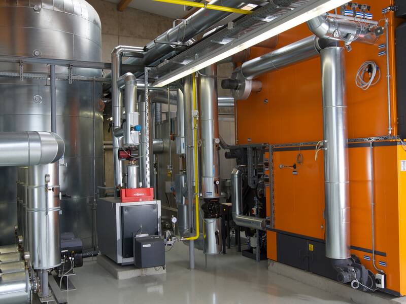 Eine Nahwärmezentrale mit großen metallischen Rohren, darunter ein auffällig orangefarbener Heizkessel.