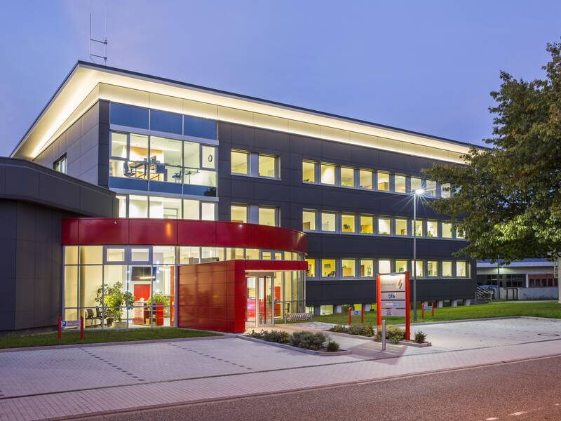 Ein modernes, dreistöckiges Bürogebäude mit großen Fenstern und einem auffälligen roten Eingangsbereich, beleuchtet in der Dämmerung.